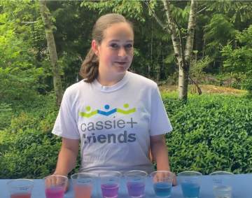 Sarah Bowers outside in a garden, with a table in front of her that has a number of cups that contain different coloured acids and bases, ranging from pink to blue. She is wearing a white T-shirt with the Cassie and Friends logo.
