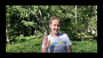 Sarah Bowers waving at the camera while being filmed for a Science at Home video. She is wearing a white T-shirt with the Cassie and Friends logo.
